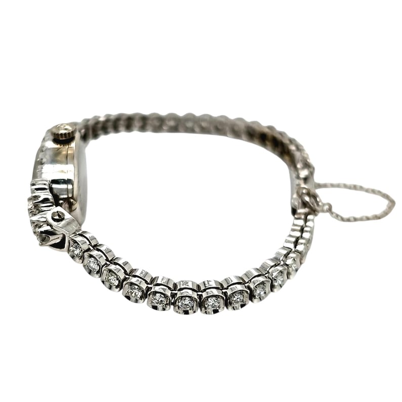May include: A silver-toned wristwatch with a delicate, linked band. The watch face is small and round, with a silver bezel. The band is adorned with small, round, clear gemstones. The watch is designed for a small wrist.