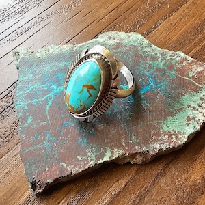 Könnte beinhalten: Ein silberner Ring mit einem Türkis-Stein, der in einer silbernen Fassung sitzt. Der Stein ist oval geformt und hat eine blaugrüne Farbe mit braunen Adern. Der Ring liegt auf einer braunen und grünen Steinoberfläche.