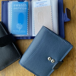May include: A blue leather wallet with a snap closure and a clear plastic insert for holding fabric swatches. The insert has a card with the text "swatch service mycolourstylist.co.uk" and a fabric swatch labeled "light summer biscuit top ten neutral".