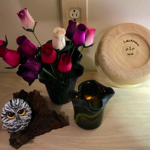 May include: A wooden bowl with the text "J. Neerman M & E 2020" inscribed on the bottom. A black vase with a bouquet of pink, purple, and white roses. A small owl painted on a rock and a black glass vase with a yellow and green design.