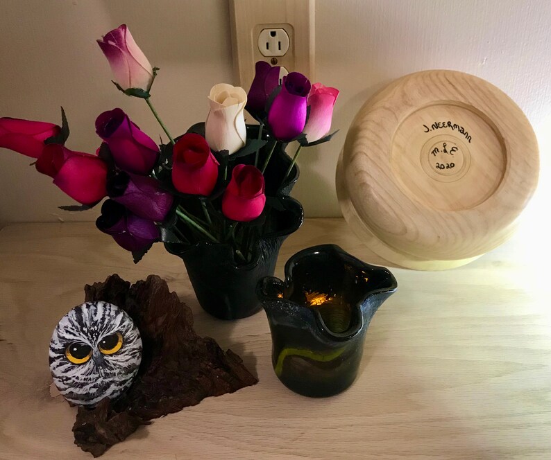 May include: A wooden bowl with the text "J. Neerman M & E 2020" inscribed on the bottom. A black vase with a bouquet of pink, purple, and white roses. A small owl painted on a rock and a black glass vase with a yellow and green design.