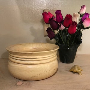 May include: A light wood bowl with a rounded bottom and a wide rim. The bowl has three raised rings around the middle. The bowl is sitting on a light wood surface. There is a small gold turtle figurine to the right of the bowl.