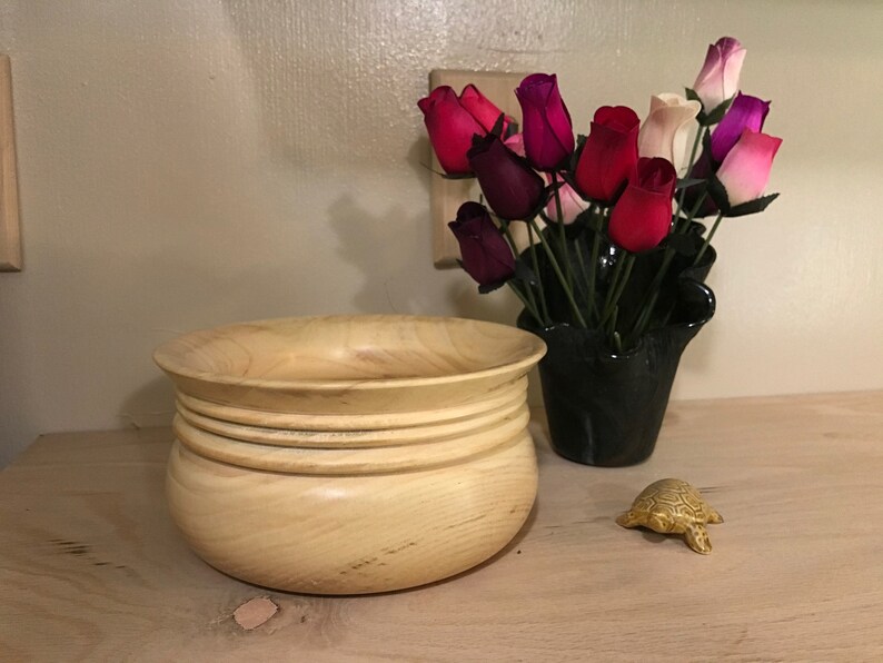 May include: A light wood bowl with a rounded bottom and a wide rim. The bowl has three raised rings around the middle. The bowl is sitting on a light wood surface. There is a small gold turtle figurine to the right of the bowl.