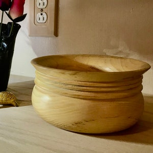May include: A large, light brown wooden bowl with a rounded bottom and a slightly flared rim. The bowl has a series of raised rings around the middle.