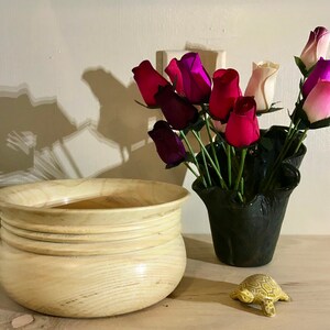 May include: A light wood bowl with a rounded bottom and four raised rings around the top edge. The bowl is sitting on a wooden surface next to a black vase filled with pink, purple, and white artificial roses. A small yellow turtle figurine is also on the surface.