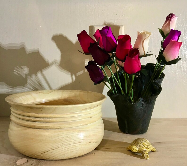 May include: A light wood bowl with a rounded bottom and four raised rings around the top edge. The bowl is sitting on a wooden surface next to a black vase filled with pink, purple, and white artificial roses. A small yellow turtle figurine is also on the surface.