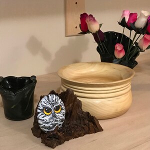 May include: A hand-painted owl rock sculpture on a wooden base. The owl has black and white feathers and yellow eyes. The wooden base is brown and has a rough texture.