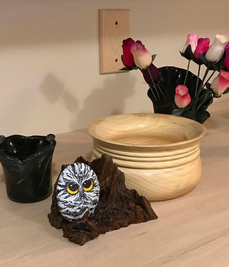 May include: A hand-painted owl rock sculpture on a wooden base. The owl has black and white feathers and yellow eyes. The wooden base is brown and has a rough texture.