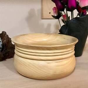 May include: A light wood bowl with a rounded bottom and a wide, shallow top. The bowl has four raised rings around the top edge.