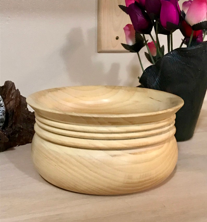May include: A light wood bowl with a rounded bottom and a wide, shallow top. The bowl has four raised rings around the top edge.