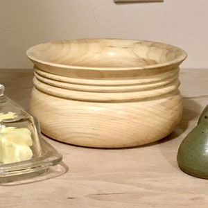 May include: A wooden bowl with a light brown finish and a series of grooves around the rim.  Two ceramic salt and pepper shakers with faces are on a wooden surface.  A glass butter dish with a clear lid is also on the surface.