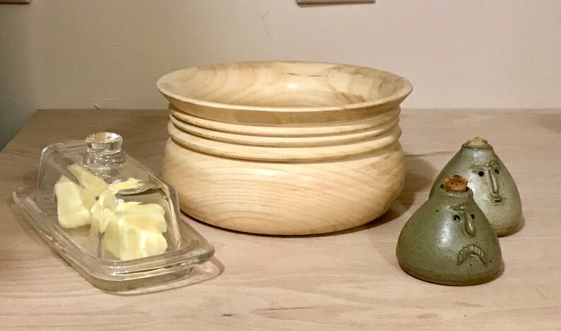 May include: A wooden bowl with a light brown finish and a series of grooves around the rim.  Two ceramic salt and pepper shakers with faces are on a wooden surface.  A glass butter dish with a clear lid is also on the surface.