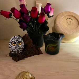 May include: A wooden bowl with the text "J. Neemann M. & C. 2020" inscribed on the bottom. A black vase with a bouquet of pink, purple, and white roses. A small owl figurine on a piece of driftwood. A small turtle figurine on a light wood surface.