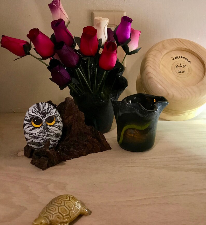 May include: A wooden bowl with the text "J. Neemann M. & C. 2020" inscribed on the bottom. A black vase with a bouquet of pink, purple, and white roses. A small owl figurine on a piece of driftwood. A small turtle figurine on a light wood surface.