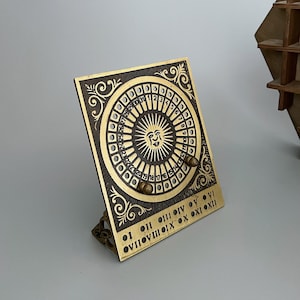 May include: A rectangular brass perpetual calendar with a dark textured background. The center features a sun face surrounded by a circular track of numbers 1 through 31 and moon phases. Below, Roman numerals I through XII are engraved in brass.