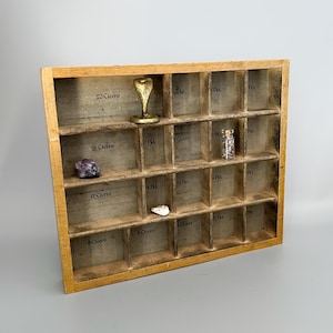 Op de afbeelding: Een rechthoekige houten letterbak met meerdere vakjes, voorzien van gedrukte labels zoals 20 Cicero en 12 Cicero. In het raster bevinden zich een koperen stempel, een kleine paarse amethistkristal, een klein glazen flesje en een kleine witte steen.