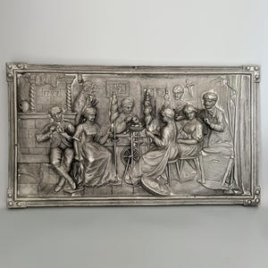 May include: A silver-coloured relief sculpture in a rectangular frame, showing a historical scene with several figures. The artwork depicts people involved in activities such as spinning and crafting. The year "1747" is visible in the upper left corner. The piece has a decorative border.