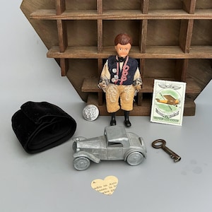 May include: A small doll wearing a dark vest and tan pants, a silver-colored metal toy car, a black velvet fabric roll, a vintage postage stamp with an airplane, a round metal coin, a small metal key, and a heart-shaped paper scrap with text.