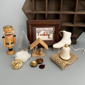 Könnte beinhalten: Eine Sammlung von Dekorationsartikeln, darunter ein hölzerner Nussknacker, ein silberner Flaschenbürstenbaum, ein gerahmter Winter-Scheunen-Druck, eine hölzerne Krippe, ein weißer Schlittschuh-Anhänger, eine Keramikglocke, eine weiße Blume und goldfarbener Schmuck.