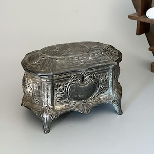 May include: An oval-shaped silver-toned metal jewellery box featuring intricate embossed floral patterns and scrollwork. The lid displays a central circular engraving, and the front includes a keyhole surrounded by detailed relief carvings on four legs.