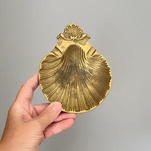 Könnte beinhalten: Eine goldfarbene Messingschale in Form einer Jakobsmuschel. Die Metalloberfläche weist ein strukturiertes, geripptes Design auf, das von der Mitte zu den gewellten Rändern verläuft, mit einem dekorativen, fächerartigen Kamm an der Oberseite.