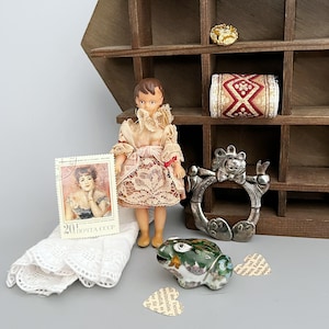 May include: A miniature doll wearing a lace dress, a vintage postage stamp featuring a woman, a silver-toned metal ring with a cat motif, a green and white ceramic frog figurine, a small roll of patterned fabric, and a gold-toned button.