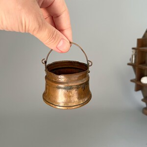May include: A small, antique copper bucket with a curved handle. The bucket has a rounded base and a slightly flared rim. The copper has a warm, aged patina, with darker areas suggesting age and use. The handle is made of a thinner copper wire.