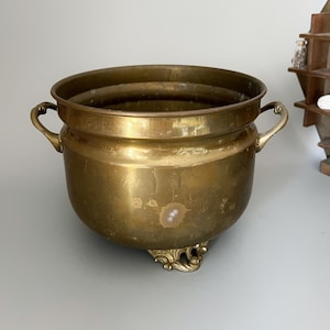 May include: A vintage brass planter with two curved handles and ornate feet. The round planter has a wide opening and a slightly textured surface. The brass has a warm, aged patina, suggesting it is an antique.