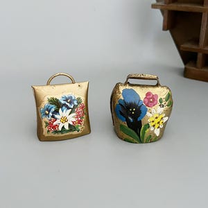 Könnte beinhalten: Zwei goldfarbene Metallglocken mit handbemalten Blumenmotiven. Die linke Glocke ist quadratisch mit einem abgerundeten Griff und einem Strauß aus blauen, weißen und roten Blumen. Die rechte Glocke ist rund mit einem rechteckigen Griff und blauen und gelben Blumen.