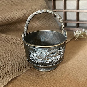 Op de afbeelding: Antieke bronskleurige emmer met een decoratief drakenmotief. De emmer heeft een stevige handgreep en een gedetailleerd reliëfontwerp rond de body. De emmer is klein en de algehele esthetiek is vintage.