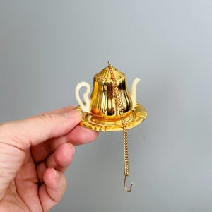 Könnte beinhalten: Ein goldfarbener Tee-Ei-Einsatz aus Metall in Form einer Miniatur-Teekanne mit cremefarbenem Griff und Ausguss. Die Teekanne steht auf einer passenden goldfarbenen Untertasse und verfügt über eine lange, dünne goldene Kette mit einem kleinen Haken.