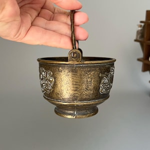 Könnte beinhalten: Ein kleiner Messing-Eimer im antiken Stil mit einem Griff. Der Eimer hat ein gealtertes, angelaufenes Aussehen und ist mit verzierten, erhabenen Reliefdesigns verziert. Der Griff ist gedreht und der Eimer hat einen abgerundeten Boden. Der Eimer hat einen Durchmesser von ca. 7,6 cm.