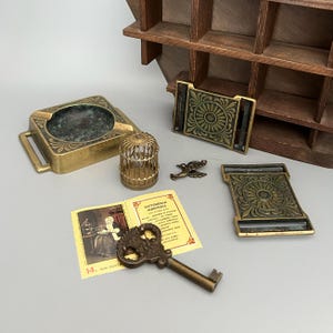 May include: A collection of brass items including a square ashtray, two rectangular belt buckles with floral patterns, a miniature birdcage, a small bird figurine, a vintage key, and a postage stamp featuring a person in a room with text.
