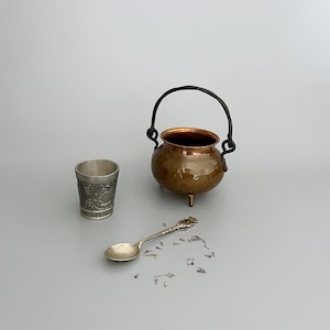 May include: A small hammered copper cauldron with a dark metal swing handle and three feet, accompanied by a silver-toned metal shot glass featuring embossed decorative patterns and a matching silver-toned metal spoon with an ornate handle.