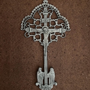 May include: Ornate silver-colored crucifix with a detailed design. The cross features the figure of Jesus Christ, with the letters INRI above. Two figures stand at the base of the cross. The background is a textured brown.