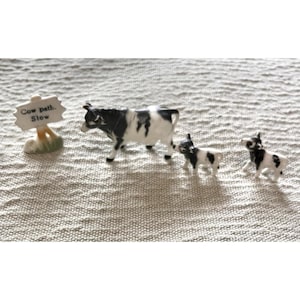May include: A set of three miniature ceramic figurines featuring a black and white cow and two smaller calves, accompanied by a small sign with a post that has the text Cow path. Slow printed in black on a white background with green accents.