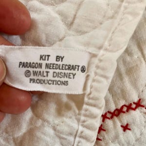 May include: Close-up of a white fabric label with the text "KIT BY PARAGON NEEDLECRAFT &copy; WALT DISNEY PRODUCTIONS". The label is attached to a quilted white fabric with red cross-stitch embroidery.