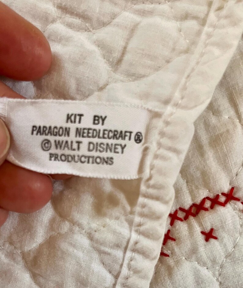 May include: Close-up of a white fabric label with the text "KIT BY PARAGON NEEDLECRAFT &copy; WALT DISNEY PRODUCTIONS". The label is attached to a quilted white fabric with red cross-stitch embroidery.