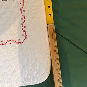 May include: A white quilted fabric with a red cross-stitch border. A wooden ruler shows the fabric is approximately 47 inches long. The fabric has a diamond pattern and a decorative stitched design.