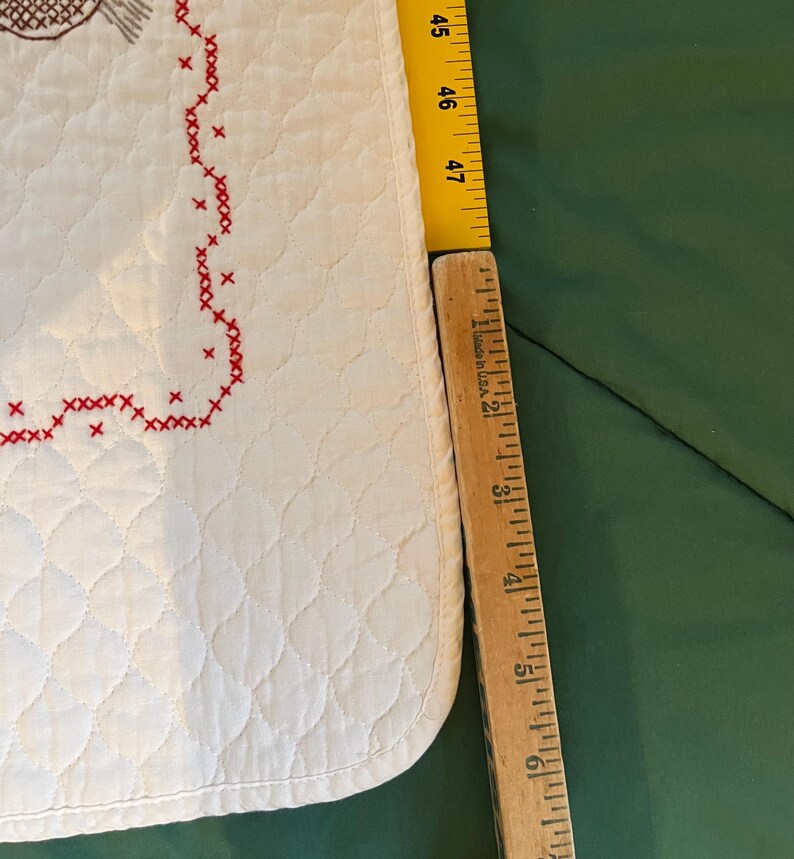 May include: A white quilted fabric with a red cross-stitch border. A wooden ruler shows the fabric is approximately 47 inches long. The fabric has a diamond pattern and a decorative stitched design.