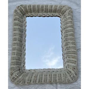 May include: A rectangular wall mirror featuring a wide, woven off-white wicker frame. The mirror has rounded corners and a smooth, reflective glass surface that shows a reflection of the sky. The wicker is tightly braided in a consistent pattern.