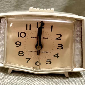 May include: A vintage General Electric alarm clock with a cream-colored face and a white plastic frame. The clock face has black numerals and hands, with the words "LIGHT DIAL" and "GENERAL ELECTRIC" printed on it. The clock is rectangular with a curved top and bottom.