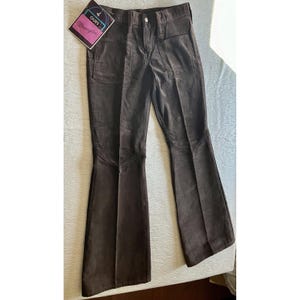 May include: Dark brown corduroy pants featuring a flared leg silhouette, a button and zipper fly closure, and two large front patch pockets. A rectangular tag with the text "OURS" and "Wrangler" is attached to the upper left side of the waistband.