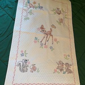May include: Cream-colored baby quilt with embroidered woodland creatures. Features a deer, owl, squirrel, skunk, birds, and rabbits. The quilt has a red stitched border and floral accents. A charming nursery item.