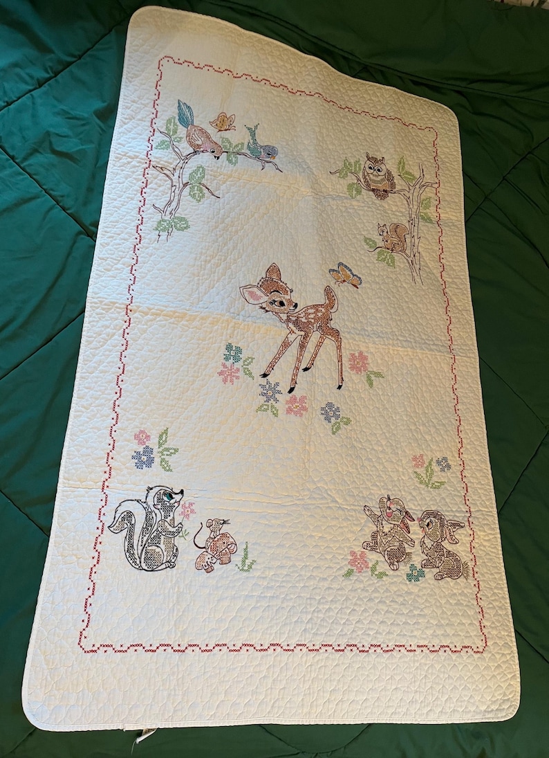 May include: Cream-colored baby quilt with embroidered woodland creatures. Features a deer, owl, squirrel, skunk, birds, and rabbits. The quilt has a red stitched border and floral accents. A charming nursery item.