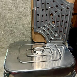 May include: A rectangular stainless steel roasting pan with a matching lid featuring a central handle. Beside it stands a flat, perforated metal rack with a cutout corner, designed for steaming or draining, showing a metallic silver finish.