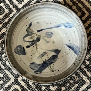 May include: A ceramic bowl with a white interior and a gray rim. The bowl is decorated with blue and gray abstract brushstrokes and speckled accents. The bowl is round and sits on a patterned surface.