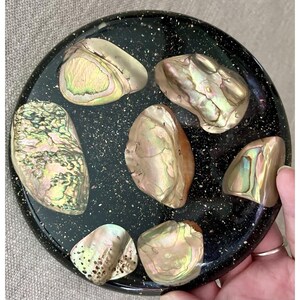 May include: A circular black tray with a speckled finish featuring six iridescent abalone shell fragments arranged on its surface. The shell pieces display natural variations in green, pink, and gold tones with a smooth, polished appearance.