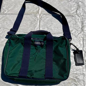May include: Green Lands' End shoulder bag with navy blue accents, including the shoulder strap and handles. The bag has a white trim and a black luggage tag. The bag has the initials "FH" and the Lands' End logo.
