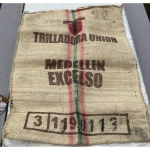 May include: A rectangular tan burlap coffee sack featuring a red and green vertical stripe. The text reads TRILLADORA UNION, MEDELLIN, EXCELSO, and the number 3 119 0 113 in a rectangular border. The woven fabric has a coarse texture.